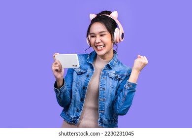 Beautiful Asian young woman in jacket jean and playing video games using joysticks with headphones on voilet background isolated.