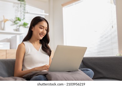 Beautiful Asian Young Woman Wear Casual Sit On Couch Using Computer Laptop Entertain With Social Media Relax Smile And Laughing.Freelance Woman Working Online Sale Marketing. Shopping Online At Home