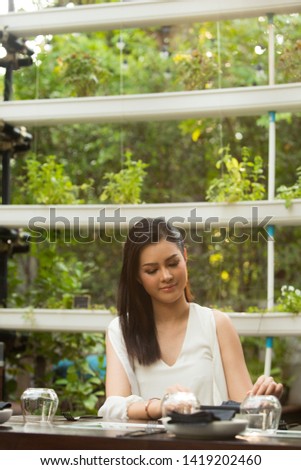 Beautiful Asian Young Woman white short dress sit in Herb garden restaurant empty glass, feel smile and happy lovely time. She wait for boyfriend to have dinner together.