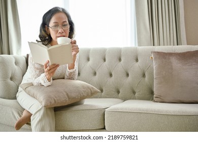 Beautiful Asian-aged woman sipping morning coffee while concentrating reading a book in her living room.