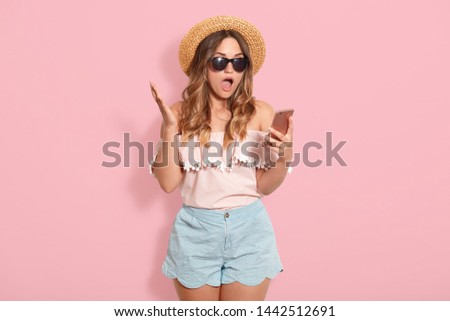 Beautiful astonished woman wearing blouse with bared shoulders, short, straw hat and sunglasses, holding smart phone in hand and posing with open mouth, recives shocking messages from friend.