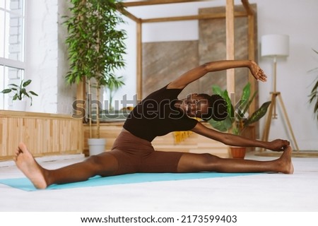 Beautiful and athletic afro american woman sitting and leaning sideways to foot, training on gymnastic mat in decorated studio. Muscle stretching, gymnastic exercises, fitness, keeping body fit.