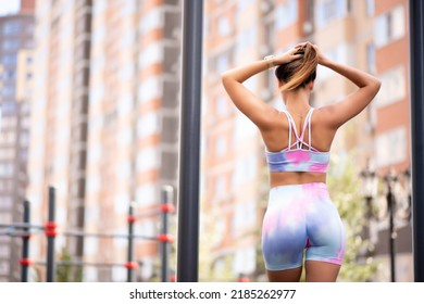 Beautiful Athletic Girl In A Pink Lavender Top And Leggings Posing On An Outdoor Sports Ground. Standing With Your Back Near The Horizontal Bar, Holding Your Hair With Your Hands