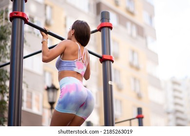 Beautiful Athletic Girl In A Pink Lavender Top And Leggings Posing On An Outdoor Sports Ground. Pull-ups On The Horizontal Bar. Hand Exercise. Motivation