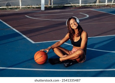 beautiful athletic middle-aged woman sitting in headphones and smiles, resting on the basketball court with a basketball next to her, tropics - Powered by Shutterstock