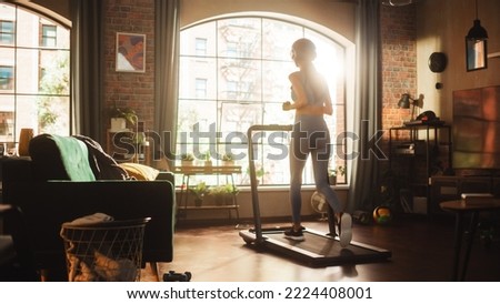 Beautiful Athletic Sports Woman Running on a Treadmill at Her Home Gym. Workout Female Athlete Training while Listening Podcast or Music in Headphones. Apartment with Window View. Side Back View