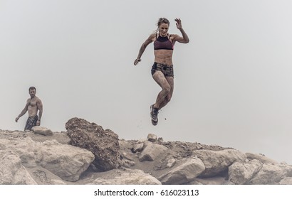 Beautiful Athletic Woman Showing Abs Jumps Of A Rock Ledge  Covered With Mud In An Extreme Sport Race At The Edge Of A Desert