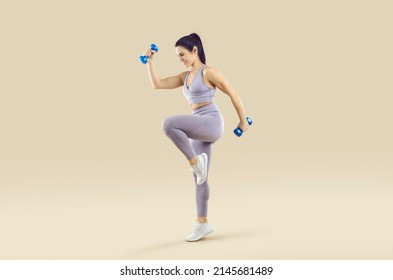 Beautiful athletic woman in sports clothes having active fitness workout. Happy fit lady in lilac crop top and leggings holding dumbbells and doing high knee exercise isolated on beige background - Powered by Shutterstock