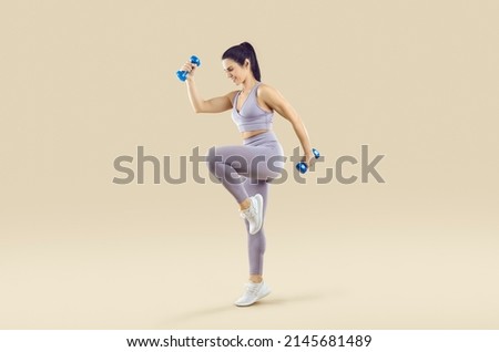 Beautiful athletic woman in sports clothes having active fitness workout. Happy fit lady in lilac crop top and leggings holding dumbbells and doing high knee exercise isolated on beige background