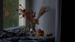 Beautiful Atmospheric Photograph Of Autumn Mood.Vase With Branches Of Orange Physalis, Pumpkins,light Garland,blanket And Book On Windowsill Near Window Wet From Rain.Autumn, Fall, Hygge Home Decor
