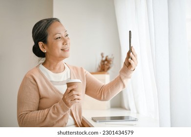 Beautiful and attractive 60s mature Asian woman taking selfie or talking on video call through her smartphone while relaxing in a cafe. - Powered by Shutterstock