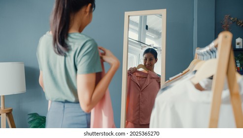 Belle et jolie femme asiatique choisissant ses vêtements sur un porte-vêtements en train de se regarder dans un miroir dans le salon de la maison. Les filles pensent quoi porter une chemise décontractée. Les femmes au style de vie se détendent à la maison.