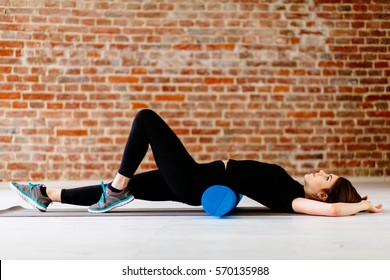  Beautiful attractive fitness woman with foam roller under back relaxing after workout on the exercising mat.Portrait of Active Tired Woman Using Blue Foam Roller in modern loft interior. 