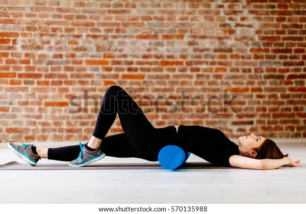  Beautiful attractive fitness woman with foam roller\
under back relaxing after workout on the exercising mat.Portrait of\
Active Tired Woman Using Blue Foam Roller in modern loft interior.\

