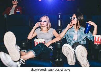 Beautiful And Attractive Girls Are Sitting In Chairs. Blonde Girl Is Talking On Thhe Phone. Her Friend Is Showing The Silence Symbol. She Wants Her To Be Quiet And Stop Talking During Watching Movie.