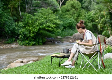 Beautiful And Attractive Millennial Asian Female Sitting On The Camping Chair, Enjoy Cooking Her Breakfast Near The River. Outdoor Activity Concept