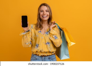 beautiful attractive smiling woman in yellow shirt and jeans holding shopping bags and using smartphone on yellow background isolated, showing phone empty screen