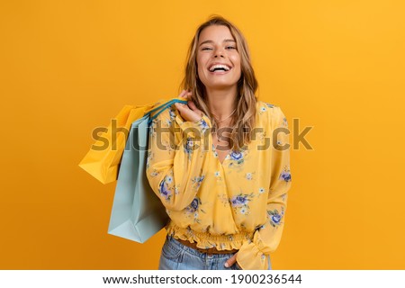 beautiful attractive smiling woman in yellow shirt and jeans holding shopping bags posing on yellow background isolated