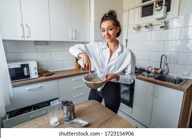 Beautiful, attractive, smiling young woman kneading dough with whisk for banana pancakes in steel dishes in a kitchen with white, light interior. Monochrome photo without bright accents