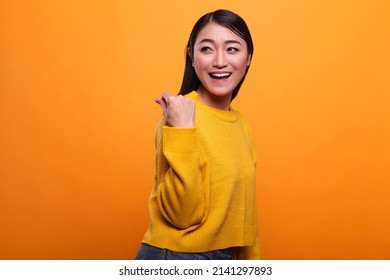 Beautiful attractive woman showing cash gesture on payday while smiling heartily on orange background. Happy positive adult person enjoying salary payout while wearing yellow vibrant sweater.