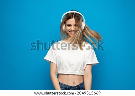 Beautiful attractive young blond woman wearing white t-shirt and glasses in white headphones listening music and dancing on blue background in studio. Relaxing and enjoying. Lifestyle.