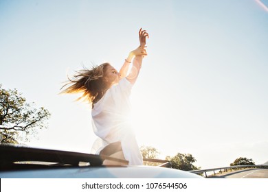 Beautiful, Attractive Young Woman With Blonde Hair And In White Tshirt Stands Up From Cabriolet Convertible Car With Sky Light, Puts Arms In Sky, Catches Wind Of Freedom And Youth, Excited And Happy