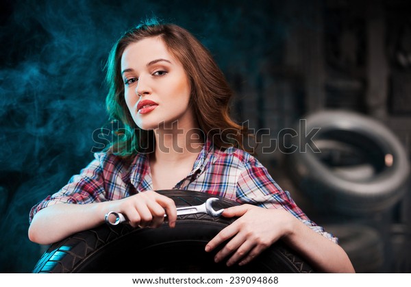 Beautiful auto mechanic. Beautiful young woman\
holding work tool and looking at camera while leaning at the car\
tire in auto repair\
shop