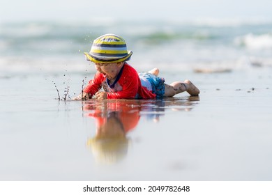 Beautiful Baby Boy Splashing Water And Crawling At A Beach. Child Is Wearing A Striped Hat. His Reflection Can Be Seen On The Water.