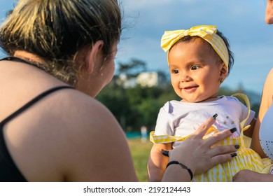 Beautiful Baby With Brown Skin, In Her Mother's Arms, Looking At Her Aunt While She Tickles Her Belly To Show Her New Baby Teeth. Beautiful Summer Day.