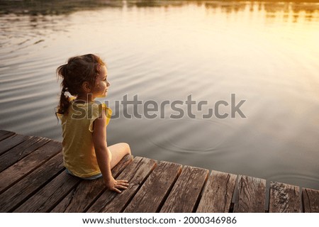 An beautiful baby girl sits on a wooden pier by the lake and enjoys the warm of a summer evening. The beautiful light of the sunset is reflected in the water of the lake.