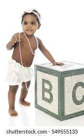 A Beautiful Baby Girl Standing With The Help Of A Giant Alphabet Block.  She's Wearing A White Hair Bow, Strands Of Beads And A Silky Diaper Cover.  On A White Background.