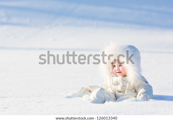 Hermoso bebé con traje blanco sentado en un campo de nieve en un día de invierno muy soleado