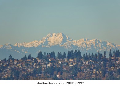 As a beautiful backdrop, Olympic Mountains, 100 miles away, rise high above Seattle hillside neighborhood filled with houses & evergreens on clear blue sky January day. Middle peaks are "The Brothers"