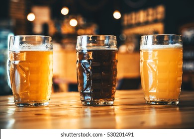 Beautiful Background Of The Oktoberfest. Glasses Of Cold Fresh White, Light And Dark Beer On The Wooden Bar Counter In The Pub. Assorted Alcohol In A Flight Ready For Tasting.