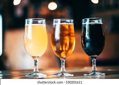 Beautiful Background Of The Oktoberfest. Glasses Of Cold Fresh White, Light And Dark Beer On The Wooden Bar Counter In The Pub. Assorted Alcohol In A Flight Ready For Tasting.