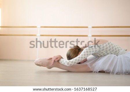 Beautiful ballerina in body and white tutu is training in a dance class. Young flexible dancer posing in pointe shoes sitting on the floor.