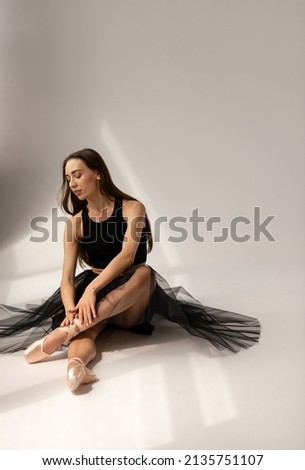 Beautiful ballerina wearing black dress sitting on floor and touching legs. Pretty ballet dancer relaxing smiling have rest in studio, on white background.