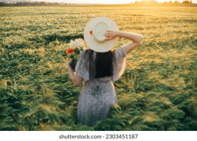 Beautiful barley field in sunset light and blurred image of woman in straw hat holding wildflowers. Evening summer countryside and gathering flowers. Atmospheric tranquil moment