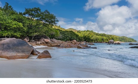 A Beautiful Beach On A Tropical Island. Picturesque Granite Boulders On The Sand. Turquoise Ocean Waves Are Foaming. A Green Hill Against A Background Of Blue Sky And Clouds Is Illuminated By The Sun.