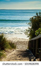 Beautiful beach pathway leading to the ocean with gentle waves lapping the shore, landscape with wooden steps and hand railing with grass and plants lining the pathway