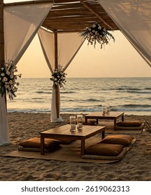 Beautiful beachside boho setup at sunset featuring a bamboo canopy with flowing white curtains, low wooden tables, cozy floor seating with leather poufs, and decorative pampas grass arrangements. 