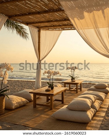 Beautiful beachside boho setup at sunset featuring a bamboo canopy with flowing white curtains, low wooden tables, cozy floor seating with leather poufs, and decorative pampas grass arrangements. 