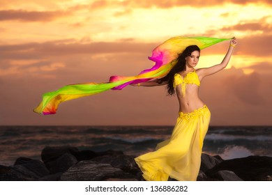 A beautiful bellydancer poses with a flowing multicolor silk veil. She is wearing a yellow costume and standing on a rocky bank in front of the Atlantic Ocean with the sun rising behind her.