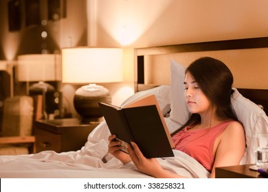 Beautiful Biracial Asian Caucasian Teen Girl Reading Book  In Bed At Night With Yellow Lamp Light On Walls