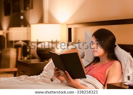 Beautiful biracial Asian Caucasian teen girl reading book  in bed at night with yellow lamp light on walls