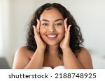 Beautiful black body positive woman touching her face and smiling at camera, making beauty treatments at home. Young african american lady enjoying selfcare