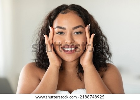 Beautiful black body positive woman touching her face and smiling at camera, making beauty treatments at home. Young african american lady enjoying selfcare