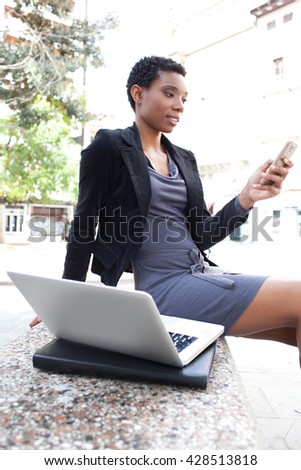 Beautiful black business woman sitting on bench in the financial city, using a smart phone and a laptop computer, smiling outdoors. Professional woman using technology, working lifestyle exterior.