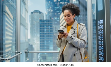 Beautiful Black Female Riding Glass Elevator to Office in Modern Business Center. Successful Manager Smile while Using Smartphone, Write Text Message, Check Social Media and Work Emails in a Lift. - Powered by Shutterstock