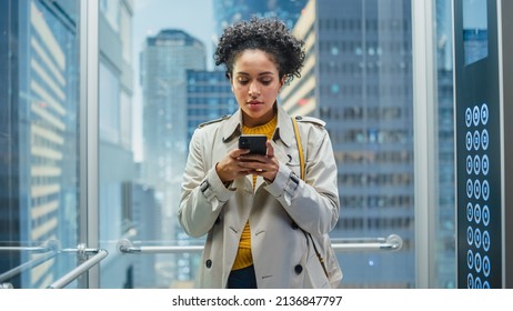 Beautiful Black Female Riding Glass Elevator to Office in Modern Business Center. Successful Manager Smile while Using Smartphone, Write Text Message, Check Social Media and Work Emails in a Lift. - Powered by Shutterstock